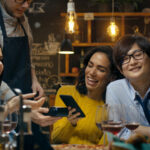 In the Bar Waiter Holds Credit Card Payment Machine and Beautiful Woman Pays for Her Order with Contactless Mobile Phone Payments System. She Has Good Time with Her Friends.